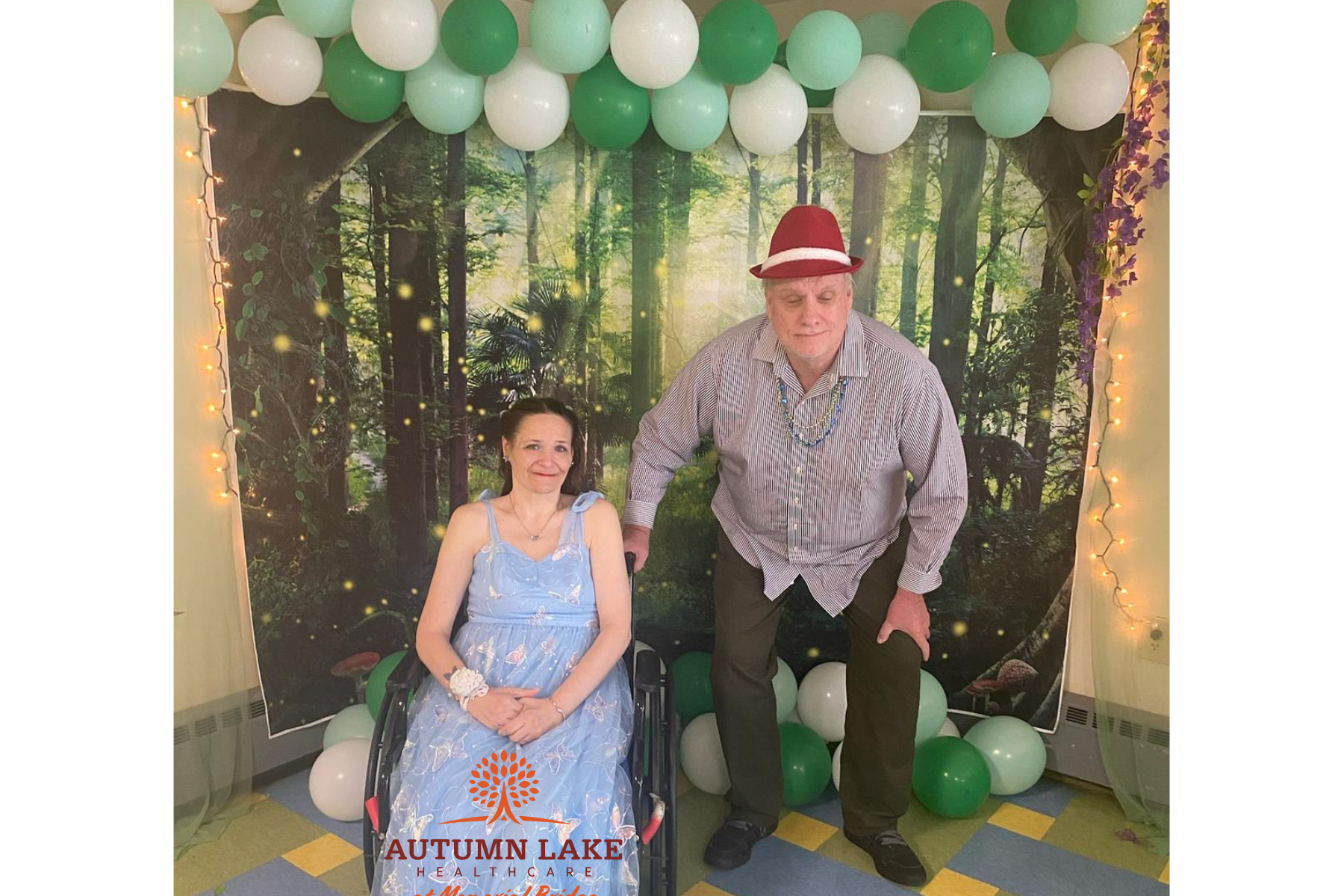 A resident in a formal blue dress and a man in a red hat pose in front of a forest-themed photo backdrop at a nursing home event.