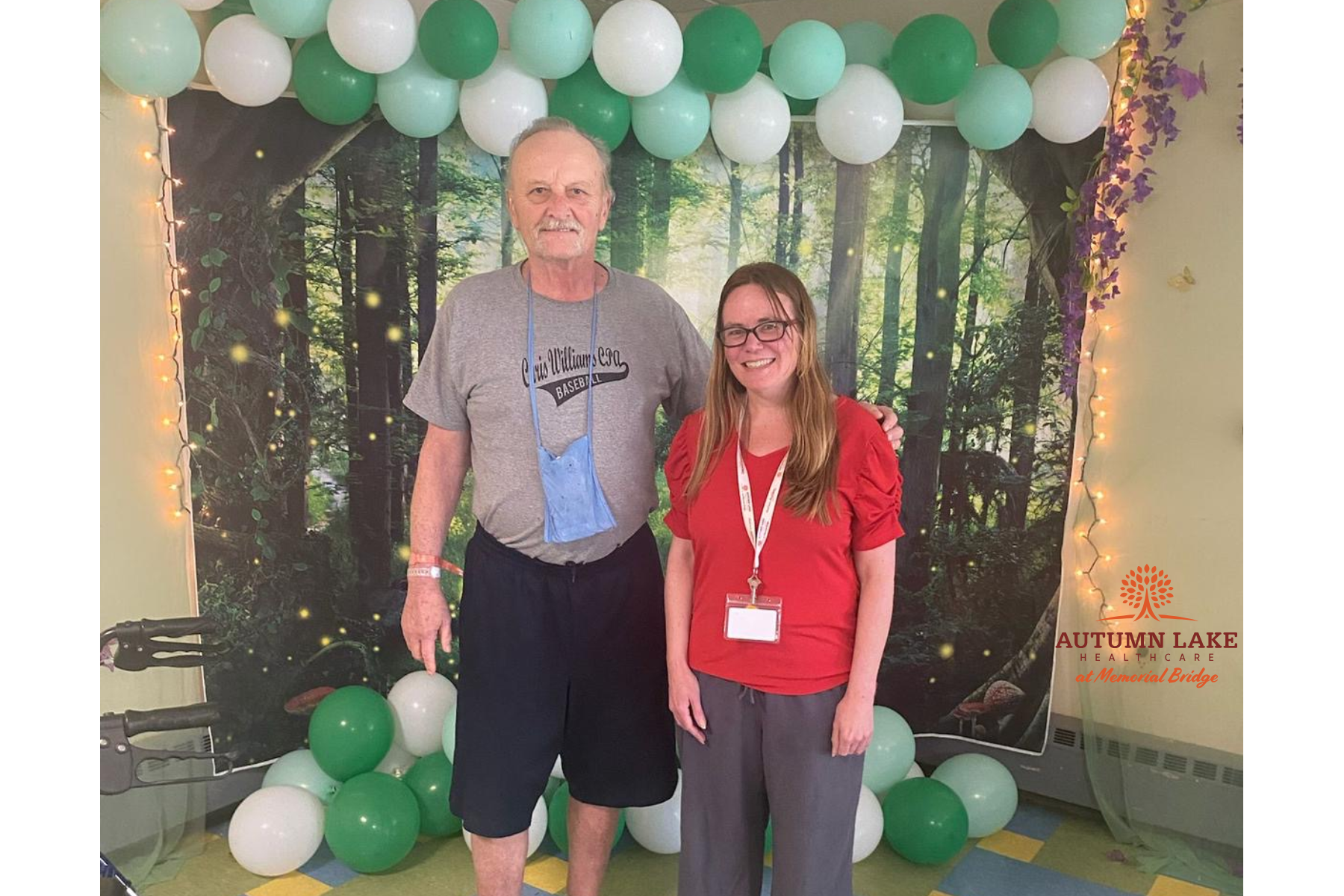 A resident and staff member pose together in front of a forest-themed photo backdrop with green and white balloons at a senior living community.