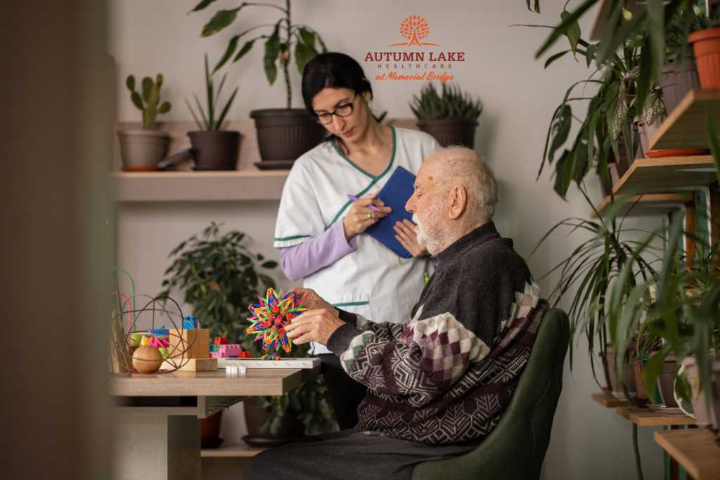 A female caregiver taking notes while an elderly man works on a colorful creative project in a plant-filled room.